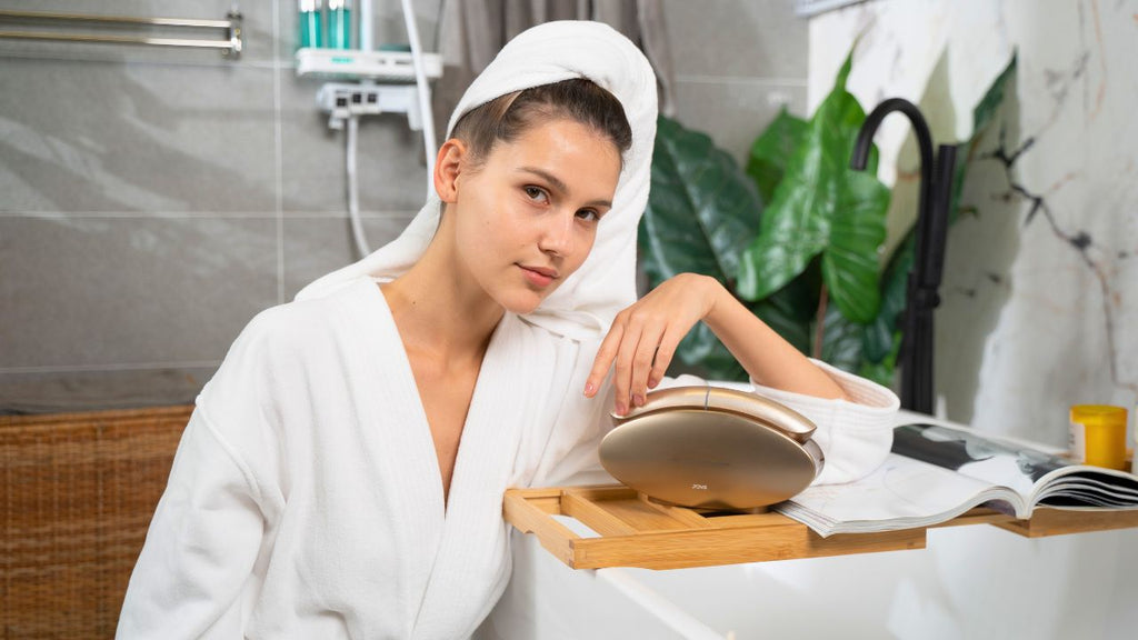 A woman in a white bathrobe with a towel on her head is looking at a microcurrent facial device on a bathtub tray.