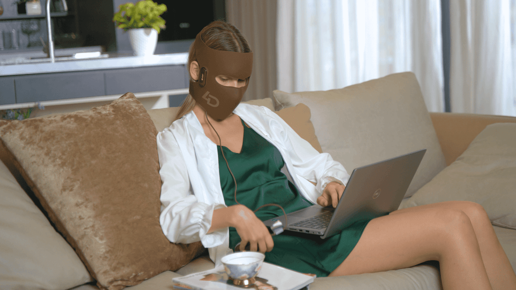 Woman using a high-tech LED laser mask while working on a laptop.