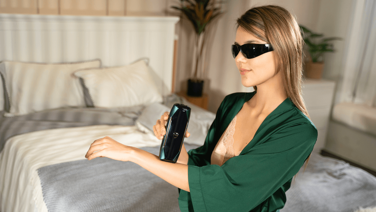 A woman in a green robe with lace details, wearing goggles, is holding a JOVS X IPL Hair Removal Device up to her forearm. The background is a softly lit bedroom with a bed and decorative pillows.