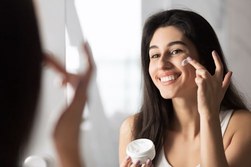 Cheerful girl applying face cream looking in mirror