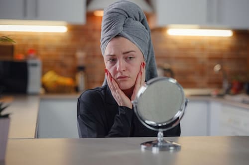 Woman with towel on her head looking at her skin with table mirror