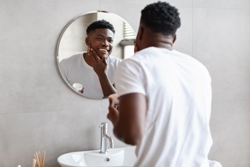 Happy black guy applying moisturizer cream on face 