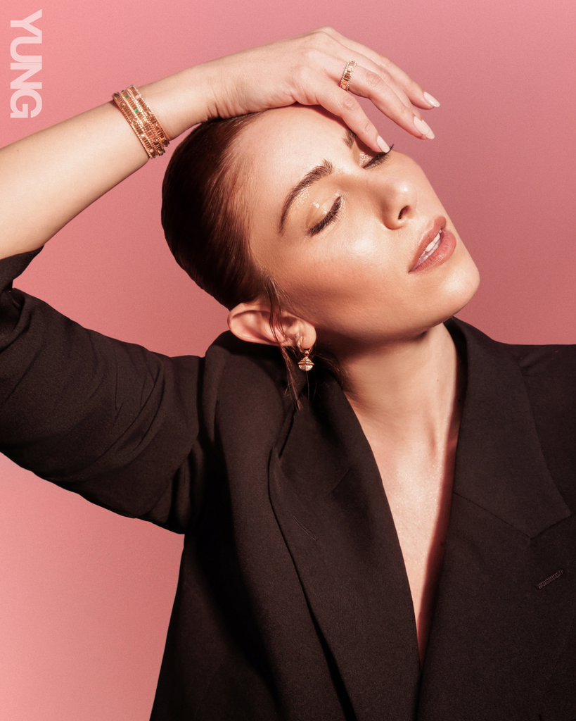 Woman wearing a black suit jacket with slicked back hair, she has her eyes closed with her hand delicately draped over her face. 