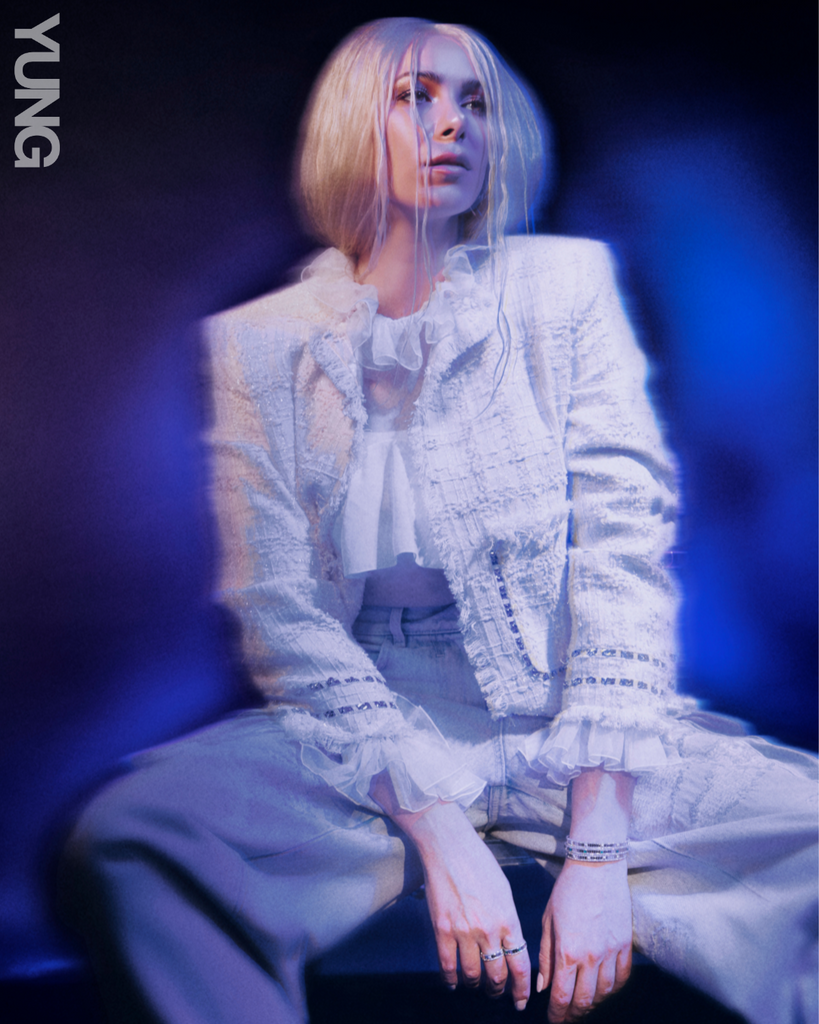 Woman with blonde hair, wearing a white suit. She is sitting her legs spread apart in a dark room with blue lighting. 