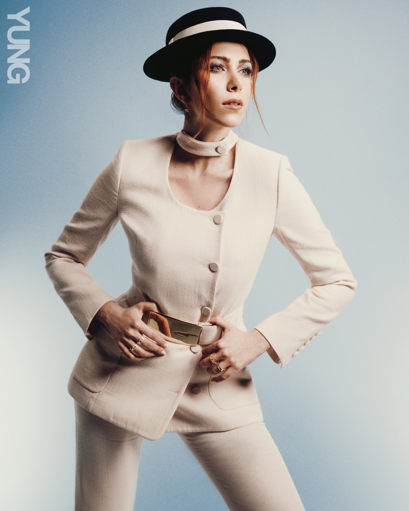 Woman wearing a black hat and a white suit. She is pushing her hips back and leaning forward her hands on her hips. She is standing in front an ombre blue backdrop.