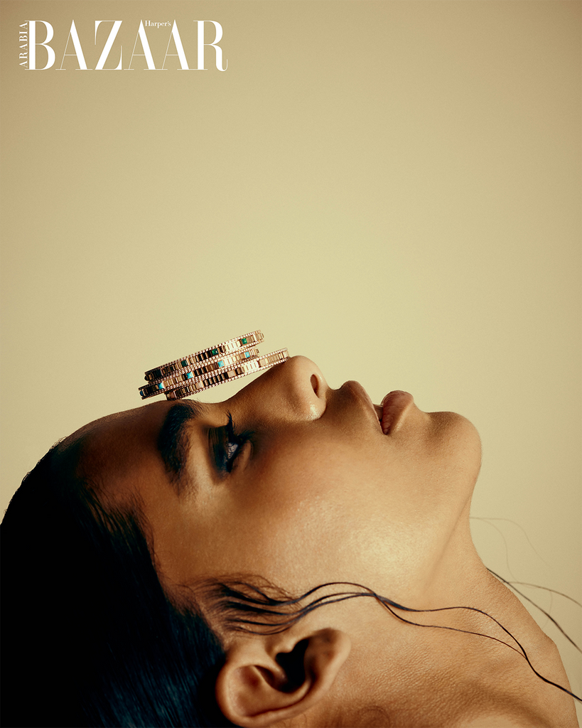 Woman with her head tilted back and bracelets balancing on her forehead and nose. She is in front of a golden yellow backdrop