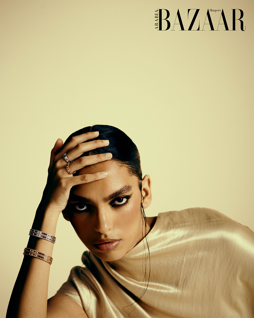 Woman looking directly at camera, resting on her elbow, and her head in her hands. She is wear bracelets and rings with her hair slicked back. She is in front of a golden yellow backdrop.