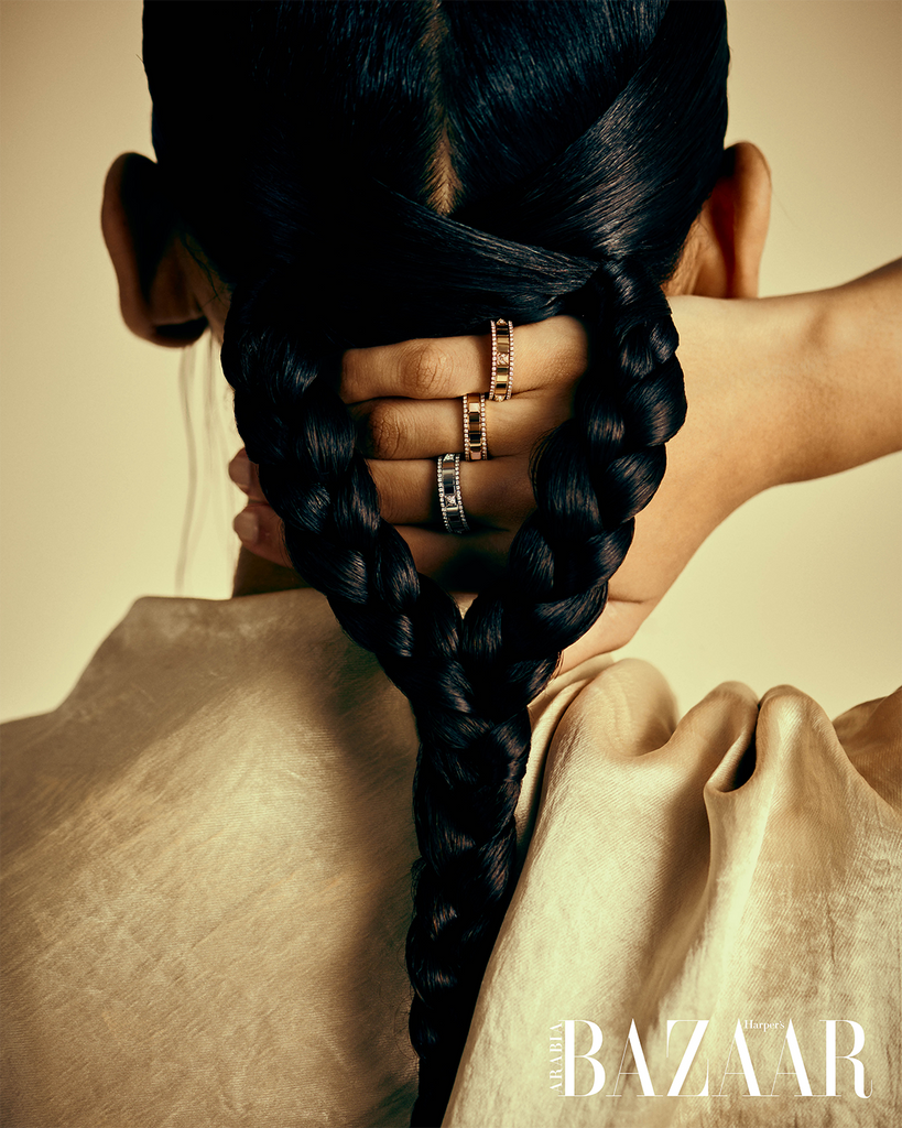 A person's hand is touching the back of their hand. They have black hair in two braids that are braided together at the end. They have their hand on the back of the head and wearing rings that you can see through the braids.