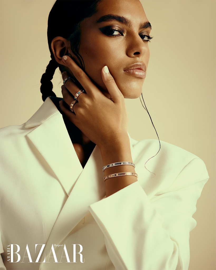 A woman in a white suit, lightly touching her face, and looking directly at the camera. She is standing in front of a golden yellow backdrop. 