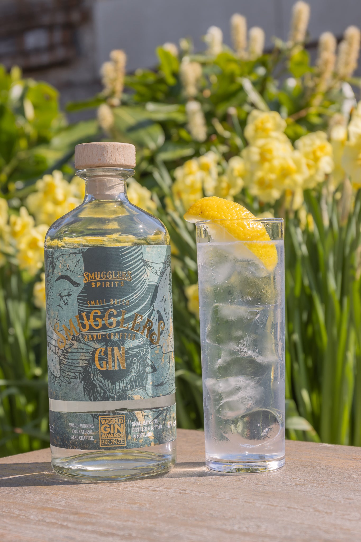 A glass of gin and tonic and a Smugglers Gin bottle sit on a table with a backdrop of daffodils