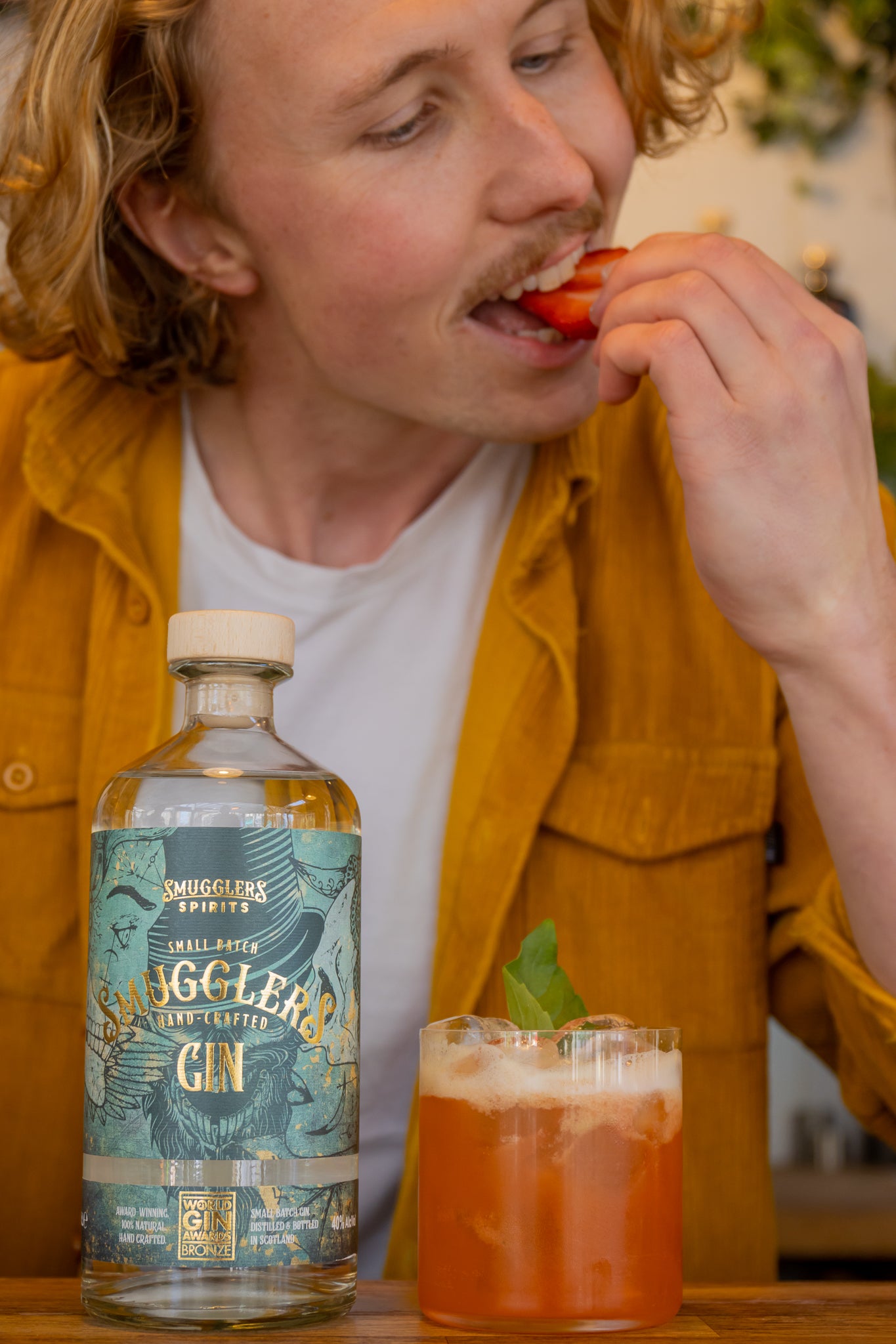Smugglers Gin bottle next to a deep red strawberry and basil cocktail, a man bites into the strawberry garnish