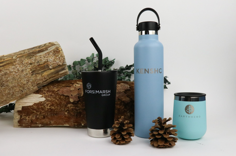 Image of a stainless steel tumbler, water bottle, and wine tumbler all custom branded with company logos. Bottles are standing in front of chopped wood and greenery