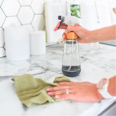 Image of hands on a kitchen counter holding a reusable glass spray bottle and a green rag