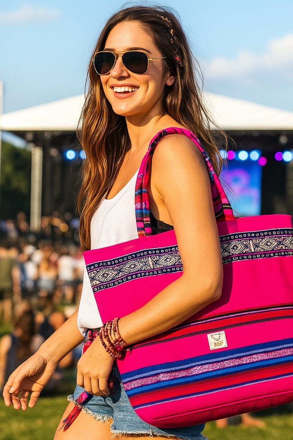 Smiling woman in sunglasses and summer clothes carrying a vibrant pink embroidered tote bag at an outdoor festival.