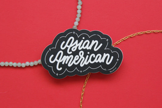A black and white JaneLi.Co sticker that says "Asian American" in cursive lettering over a red background with a string of green jade and a thin gold chain. 