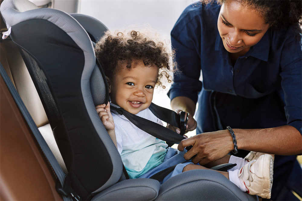 a happy little boy looking at camera while his mother buckling him in a car seat