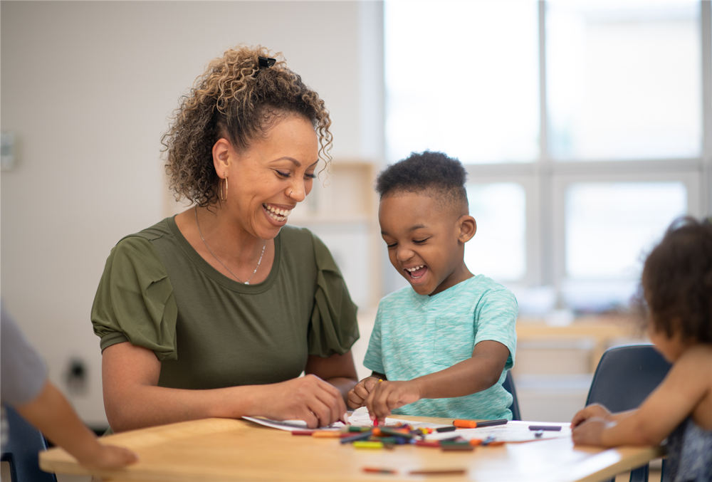 A preschool teacher and her student are colouring together at a desk