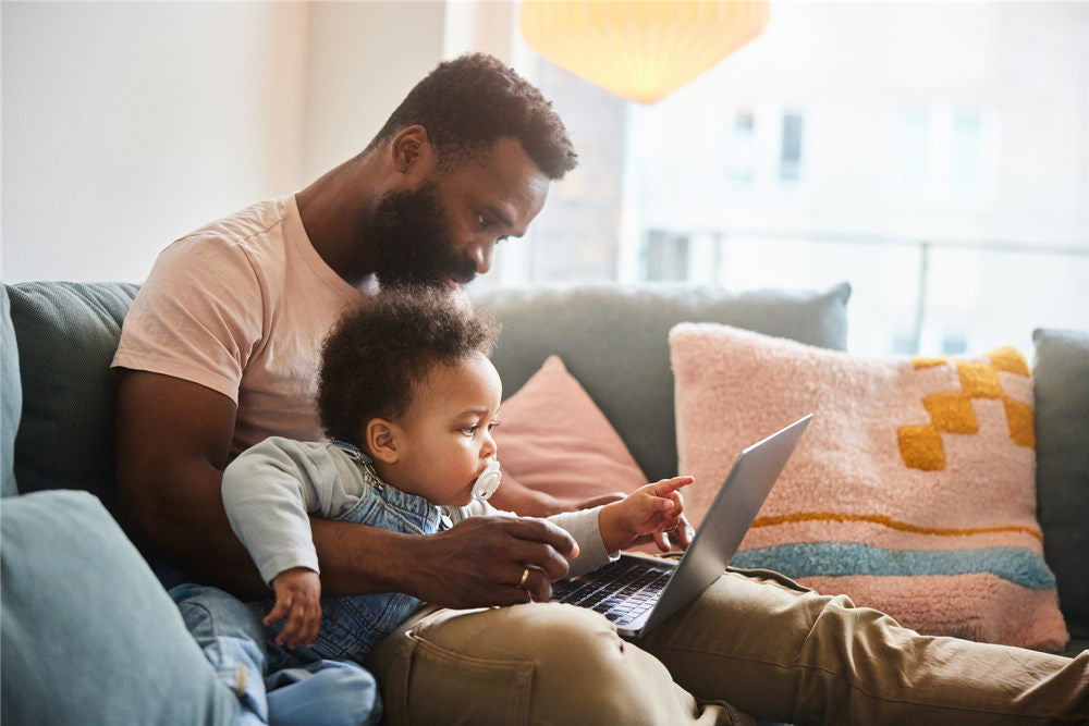 Loving father working on a laptop while sitting on a living room sofa at home with his curious baby boy on his lap