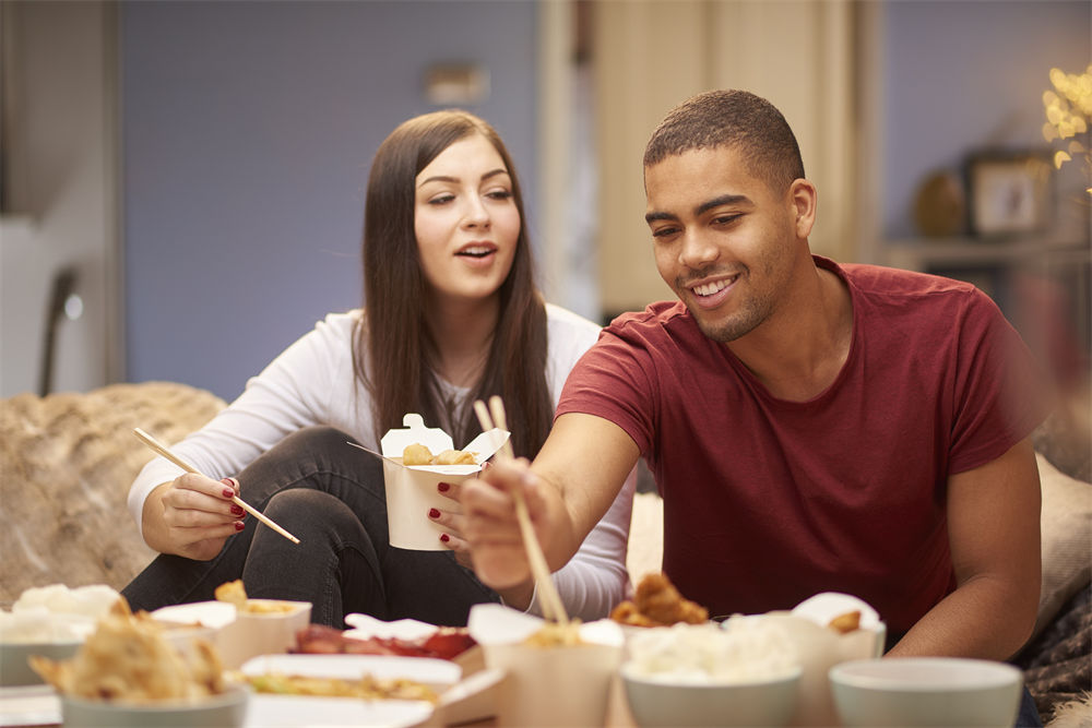 a young couple sit on the sofa and share a chinese take away meal, chatting and laughing