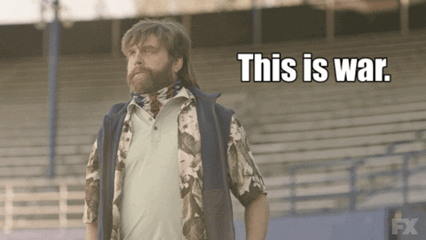GIF of a man with a mullet and beard in a polo and camo shirt standing in a stadium while stating "this is war."