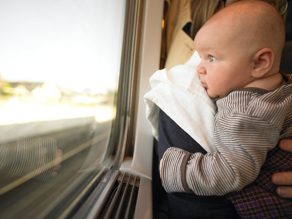 A baby gazes out the window of a train as their parent holds them.