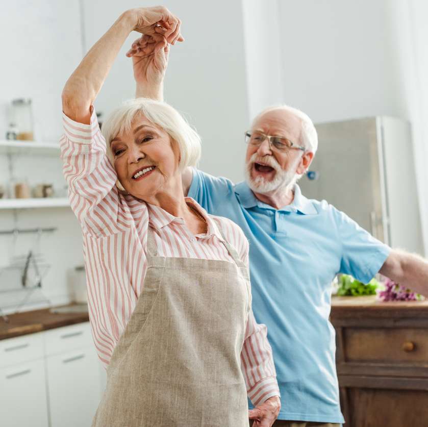Older male and woman adult dancing in the kitchen, smiling.