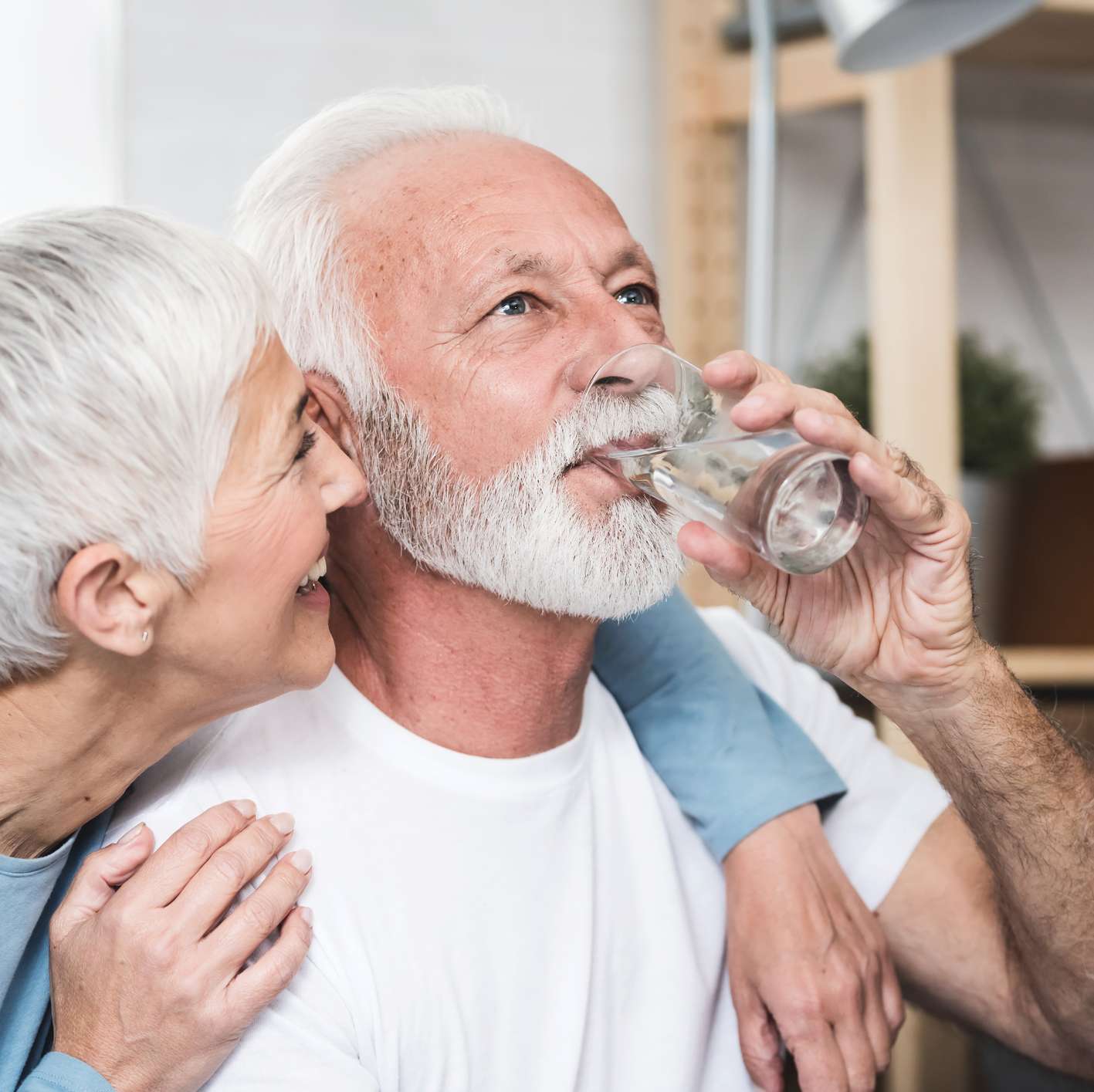 Older man drinking a glass of water with smiling wife's left arm wrapped around him.