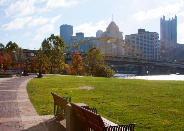 Grassy lawn by a riverbank in Pittsburgh with a yellow bridge and high rise buildings in the backdrop