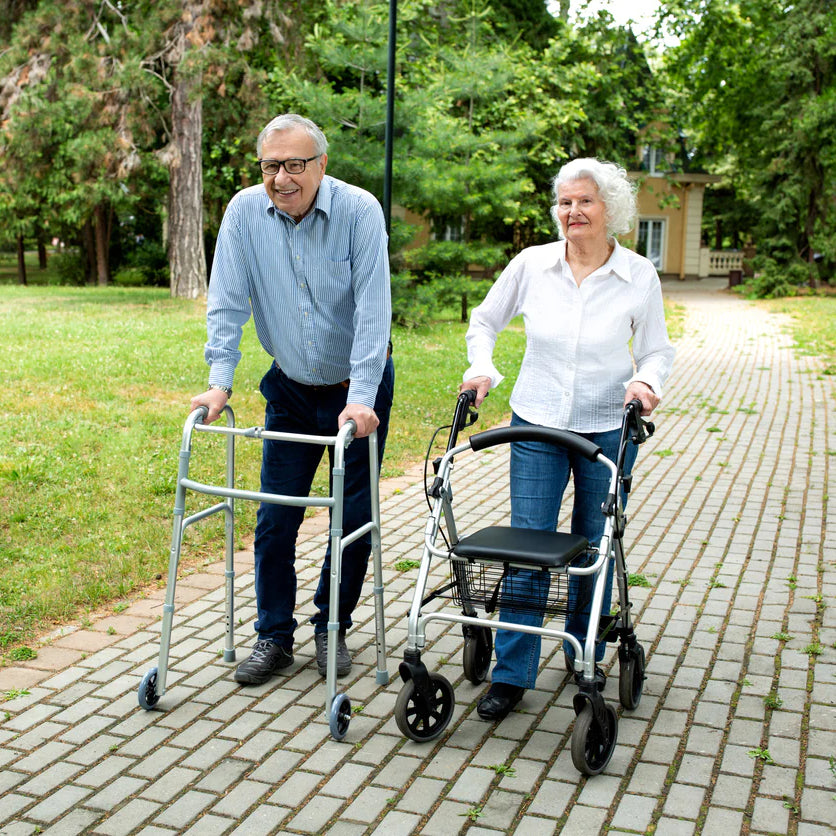 A man and a woman walk on a path outside with different types of walkers.