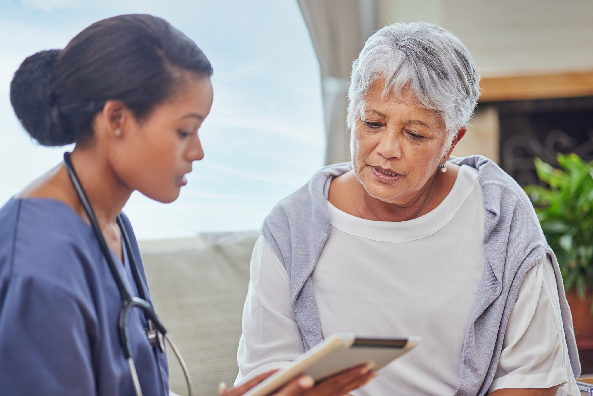 A hispanic senior woman in on a cosy sofa and her female nurse in the old age home and using a digital tablet. Mixed race young nurse and her patient talking in the lounge