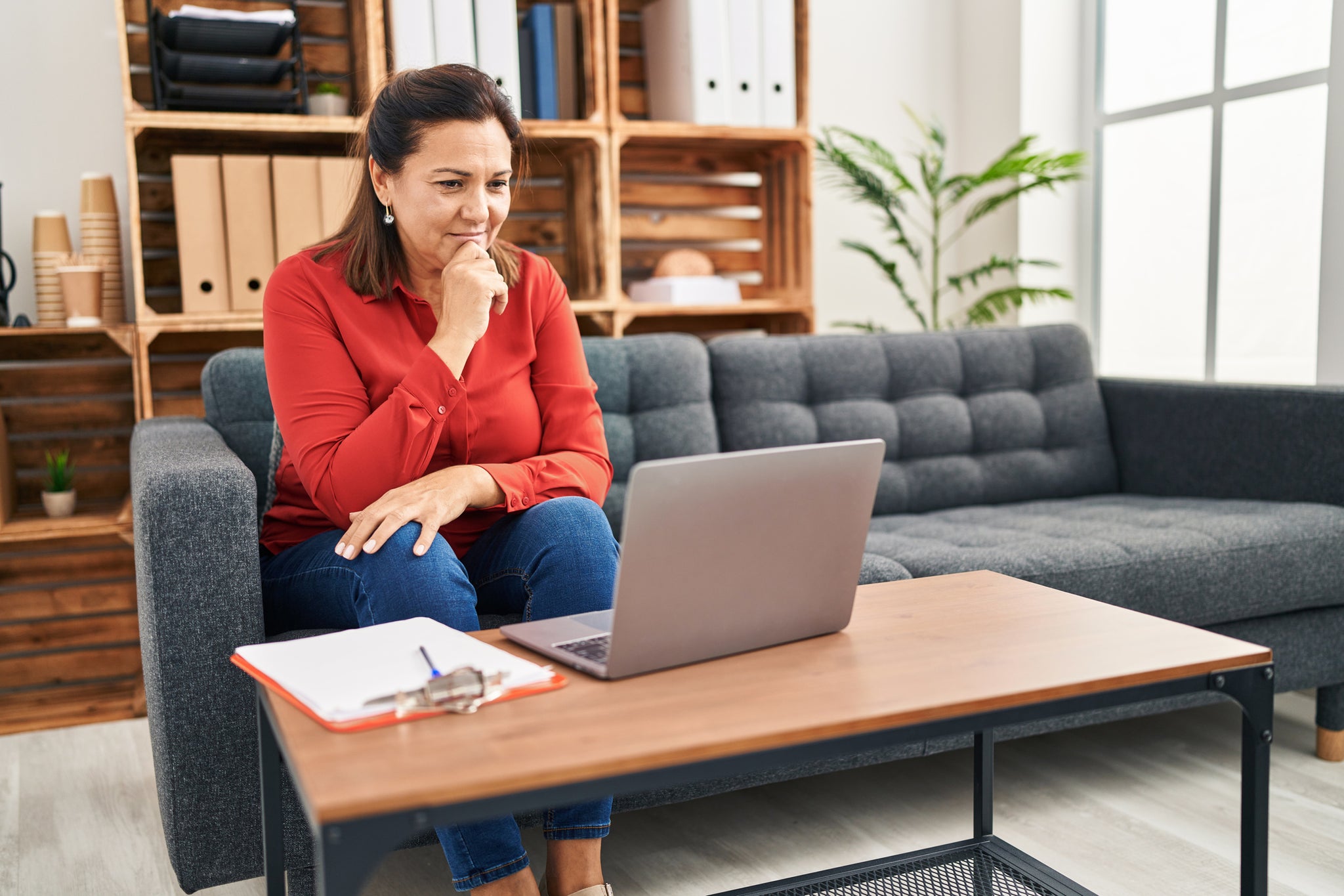 A woman looks at a computer while sitting on a couch at home.