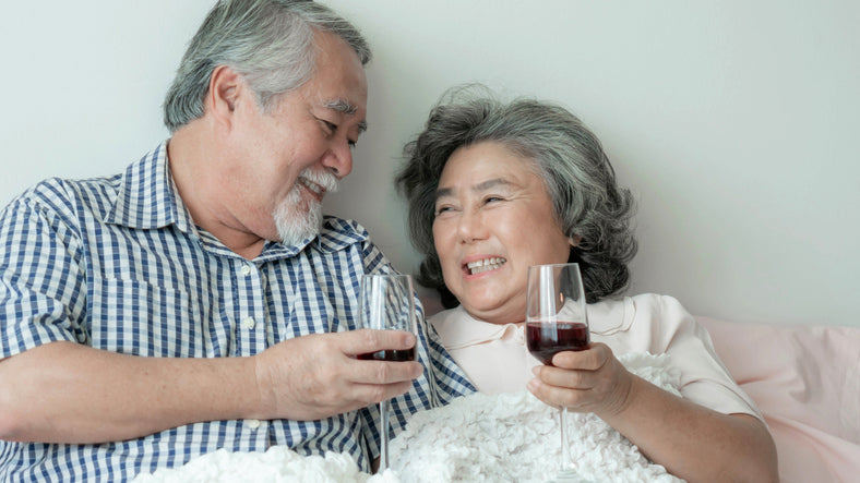A couple lays in bed smiling with glasses of red wine in their hands.