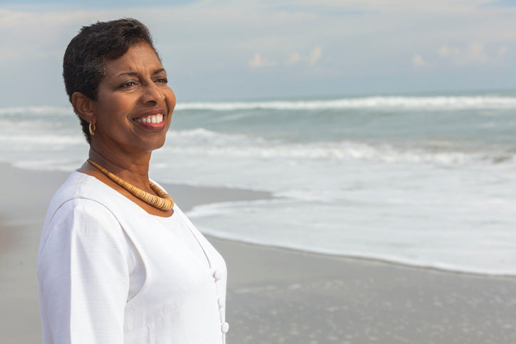 A woman stands on the beach looking out over the ocean smiling peacefully.