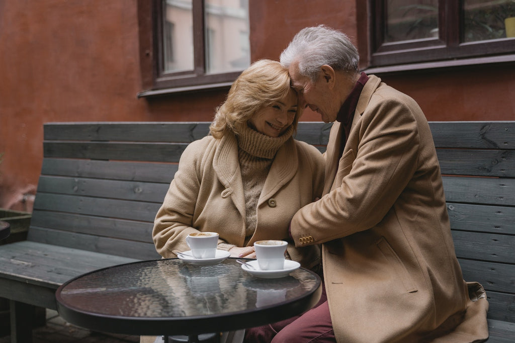 A couple sits at an outdoor table at a cafe with their heads pressed together.