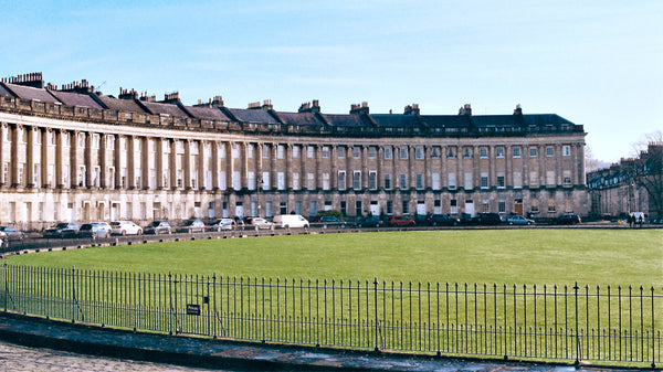 A photo of the Royal Crescent building, across its well manicured grass lawn in Bath, England. The building is a fine example of Georgian architecture.