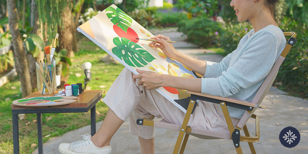 Woman painting a canvas sitted on a chair outside smiling