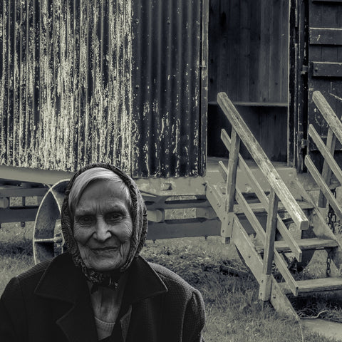 black and white photo of an old lady wearinga  headscarf in front of a gypsy caravan