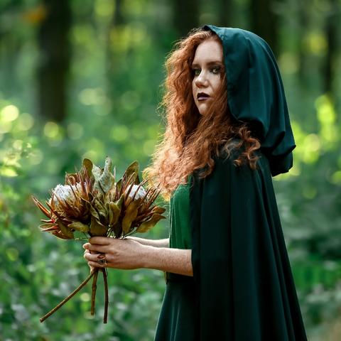 woman in a green cloak standing in the forest holding a bunch of flowers