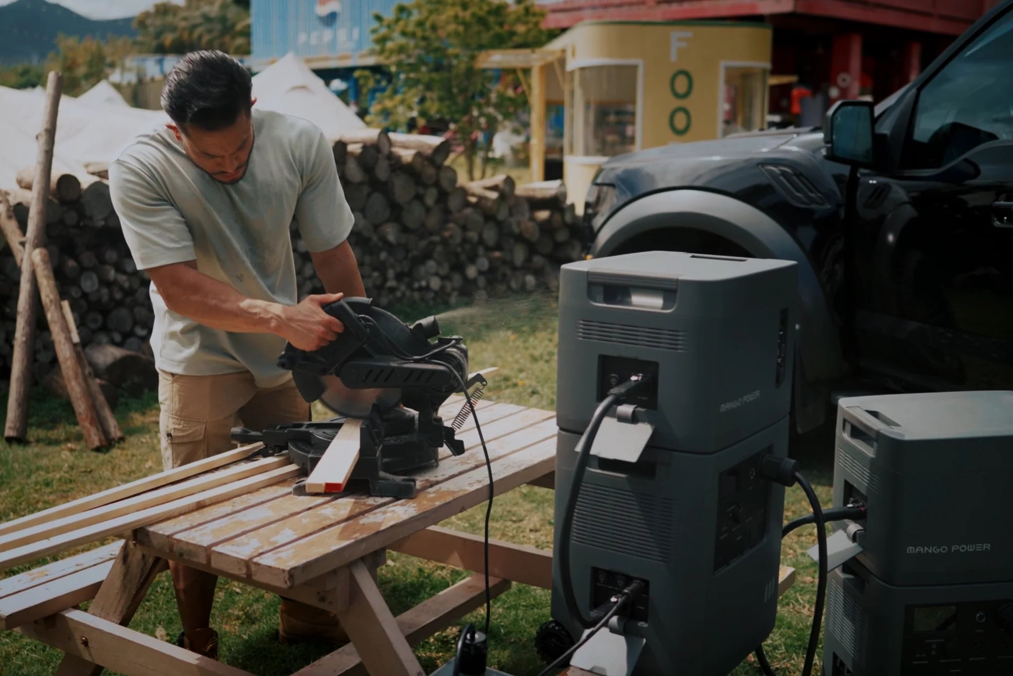 A man uses an electric saw to cut wooden planks on a picnic table outdoors, powered by the Mango Power E 6,000W 7,000Wh 240V Portable Power Station Kit with mSocket Pro from Mango Power, beside a black vehicle. Firewood stacks and buildings are in the background.