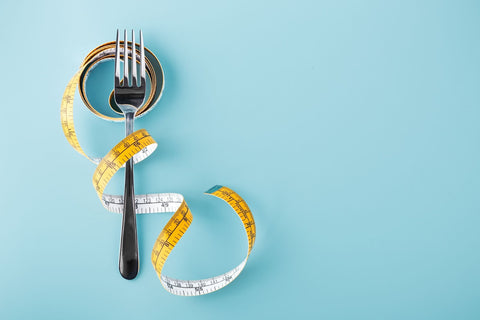 A measuring tape rests on a blue background alongside a fork and spoon, symbolizing dieting and healthy eating