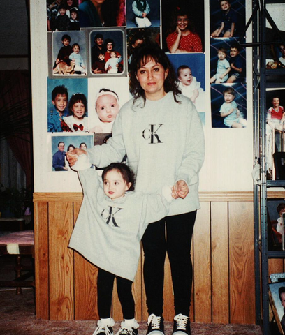 A two year old Laice holds her mothers hands in front of a wall covered in family photos. They are wearing matching outfits- grey Calvin Klein sweaters, black leggings, and black and white converse