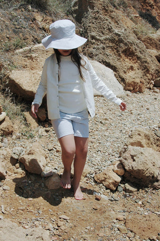 brown hair girl in blue hat, blue shorts and white top rocky beach backdrop