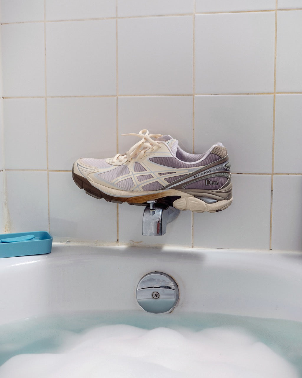 Dime x ASICS GT-2160 shoe in unique printed design, balancing on a silver faucet above a bubble-filled bathtub, representing the refreshing and innovative take on the classic ASICS silhouette.