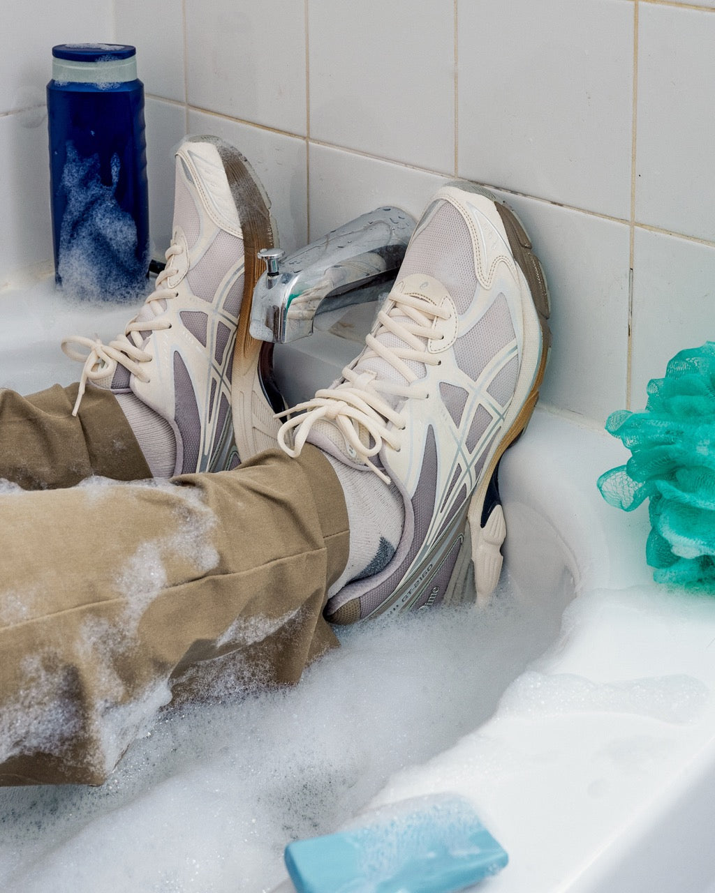 Individual fully dressed in clothes and wearing Dime x ASICS GT-2160 shoes, sitting immersed in a bathtub, playfully highlighting the sneaker's unique appeal and creative collaboration.