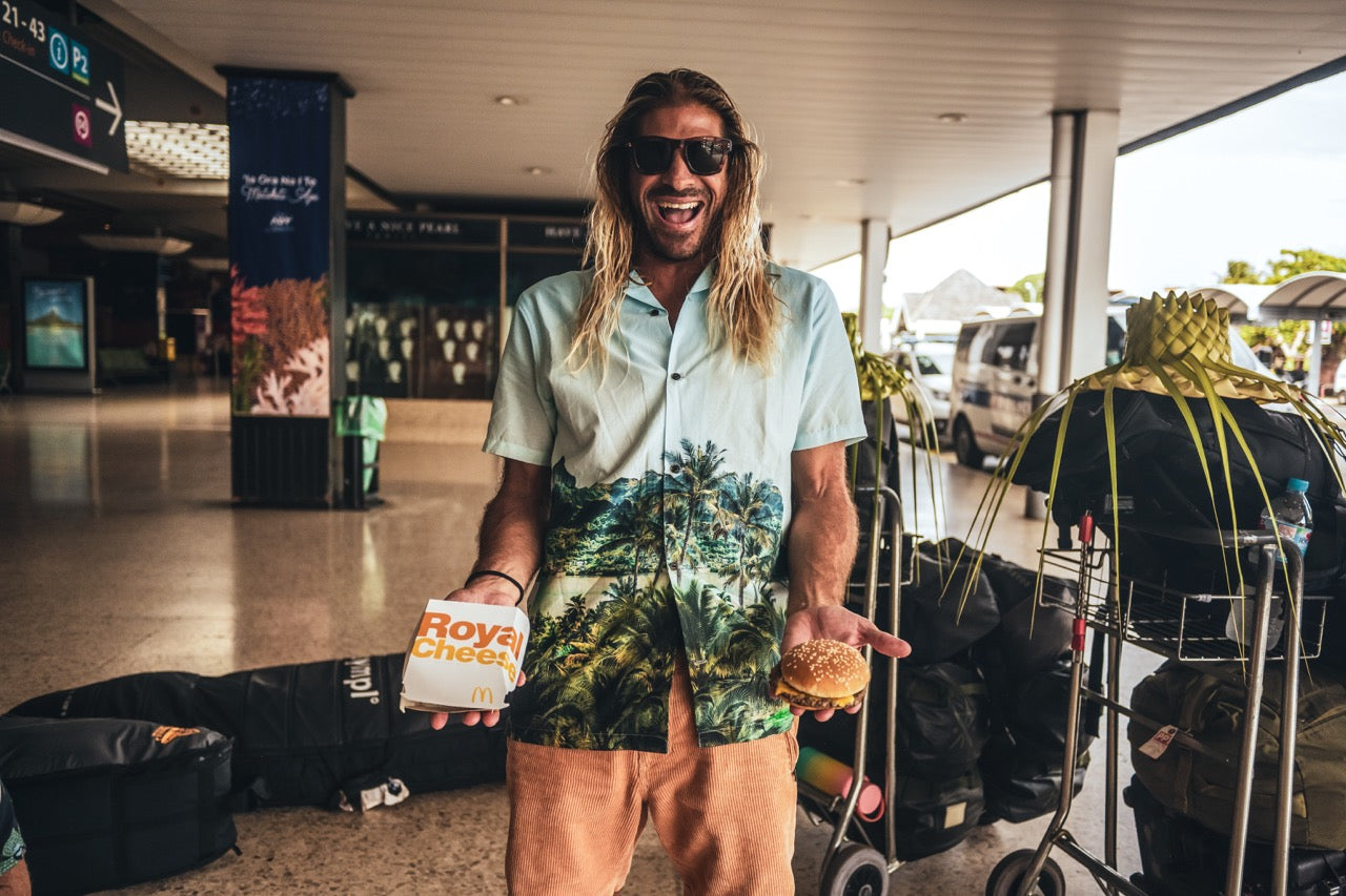 Stoked man in Tahiti's airport, confidently sporting the Roark Gonzo Camp Collar Shirt - Hinano Otemanu Light Blue, triumphantly holding a 'Royal with Cheese' with a backdrop of the bustling travel hub.