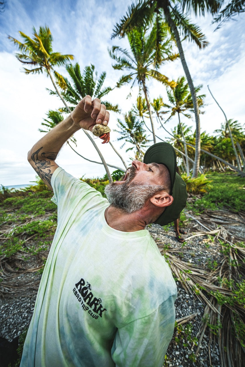 Adventurer holding a hermit crab in Tahiti wearing the Roark Road Trip Club Tie Dye Wash Premium Tee