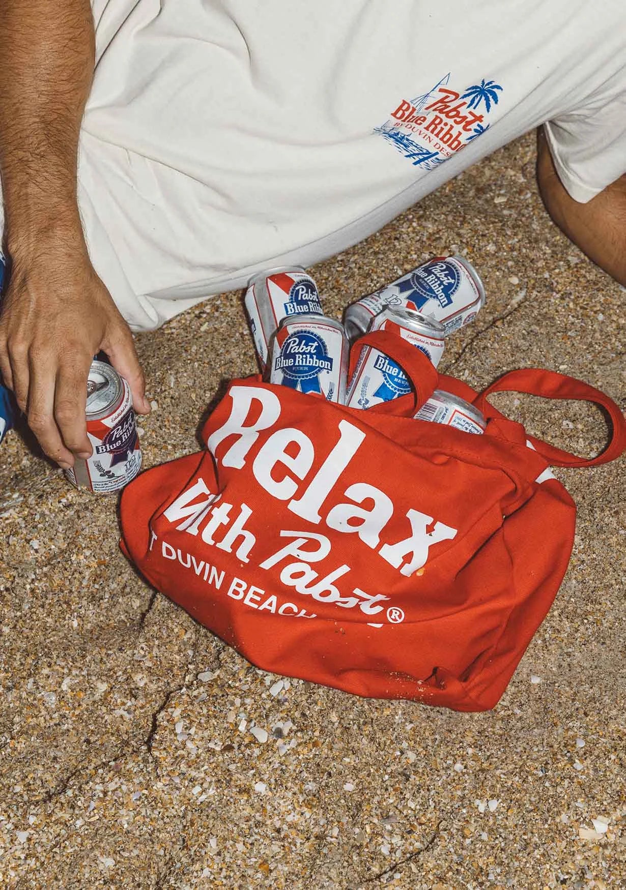 Image of a guy laying on the sand with the PBR x Duvin Tote Bag in Red