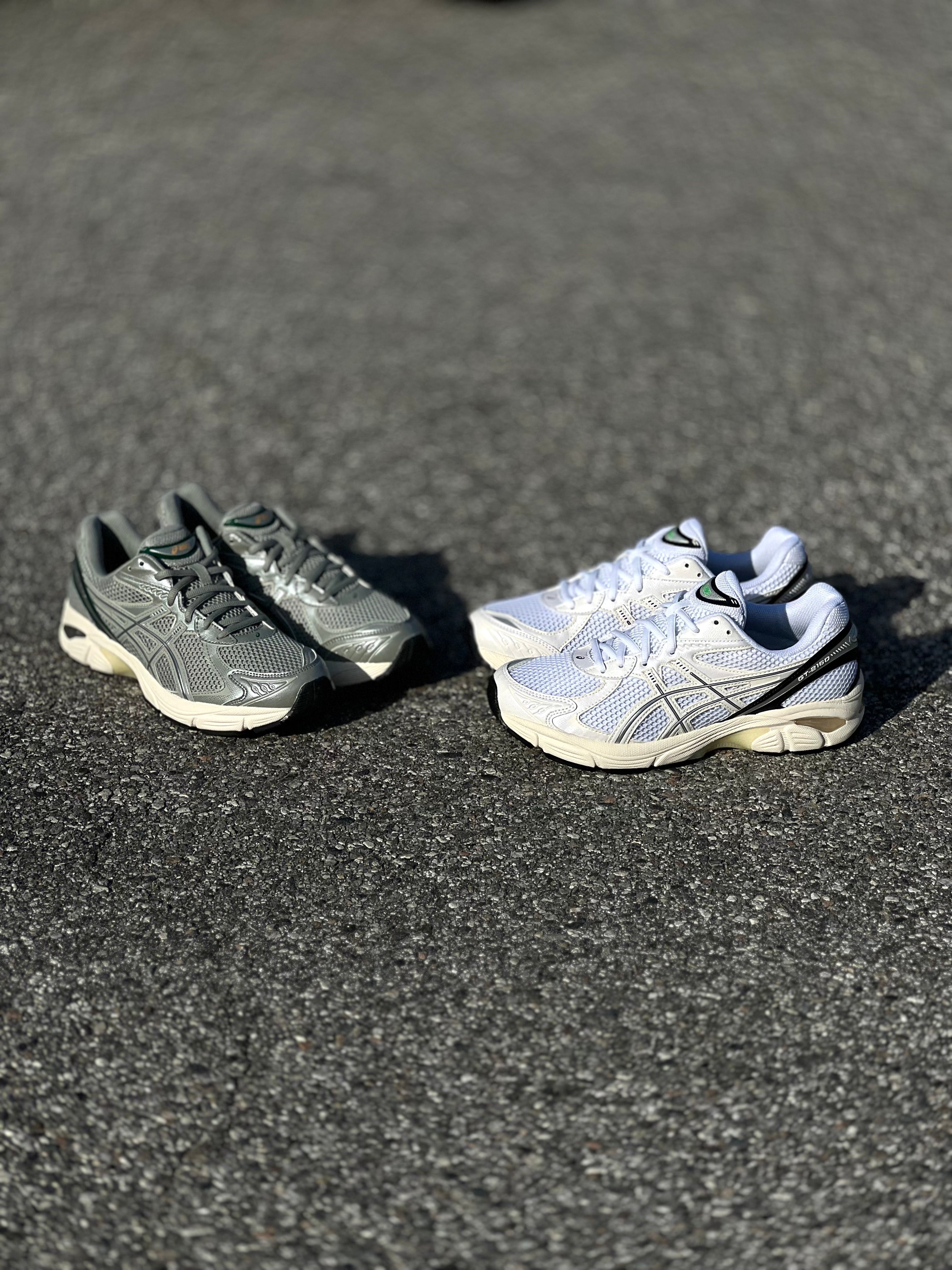 ASICS GT-2160 Seal Grey/Jewel Green and White/Black sneakers displayed on concrete at Drift House Cocoa Beach parking lot.