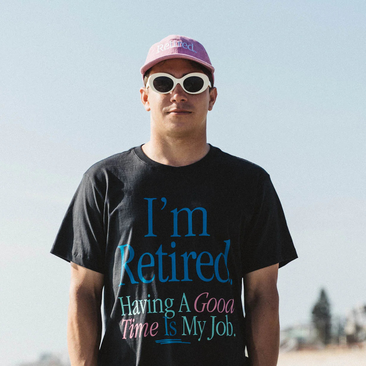 Man standing in the ocean, confidently wearing Duvin's black 'Retired' tee and pink 'Retired' hat.
