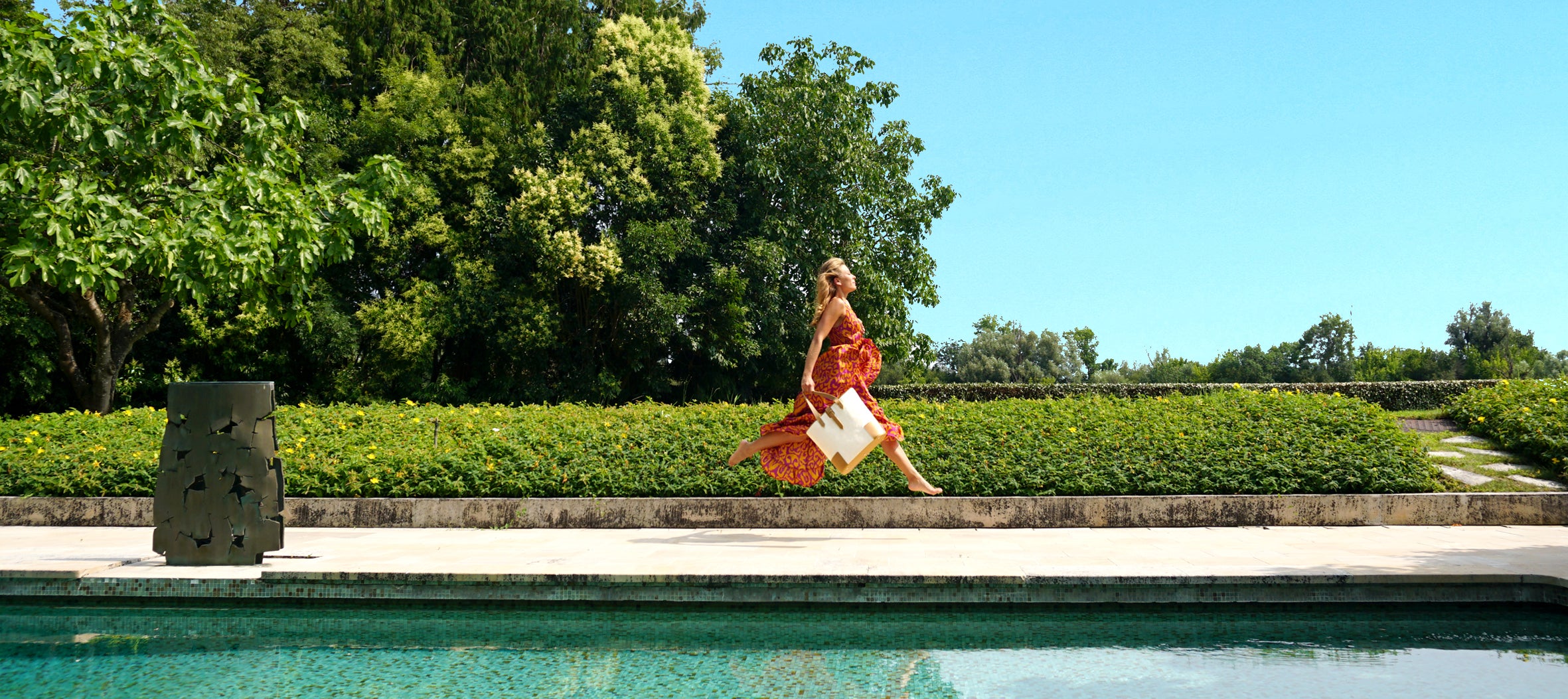 Blonde girl with orange dress and Maison Marrain DeuxMag Totebag jumping in the air next to a pool